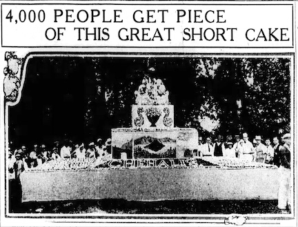 Picture of people standing behind a long banquet table holding a large cake. The cake towers over the people.